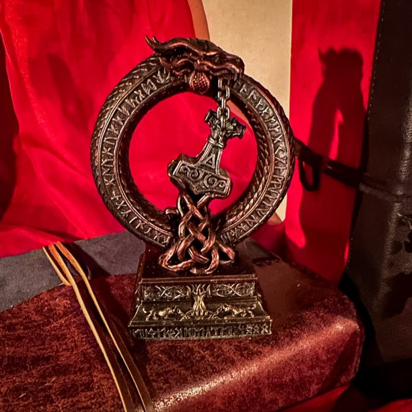 Sculpture nordique Mjölnir en résine posée sur un grimoire ancien relié en cuir dans une lumière chaude.